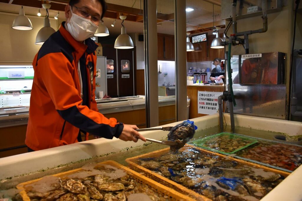 体験型食事処「炭焼　炙屋」の店内とカキを手にする田辺さん