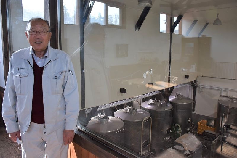 ガラス越しに見える醸造タンクなどワイン醸造設備とその前に立つ倉内武美社長