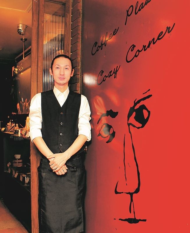 珈琲プラザ コージーコーナーの入り口にある、常連客が描いた顔の絵の前に立つ店長の後藤正吉さん