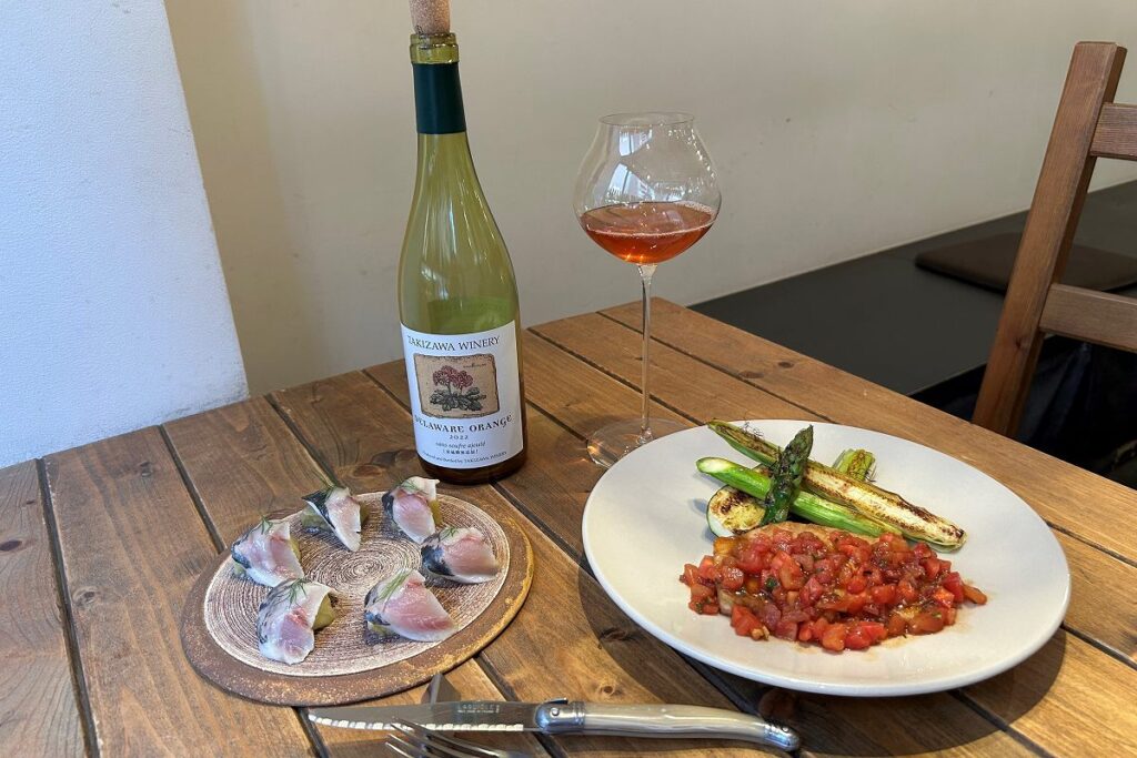 「ステム」が提供しているアジと焼きなすのマリネ(左)とメカジキのフルーツトマトソース、グラスに注がれた「タキザワワイナリー デラウェアオレンジ」とボトル