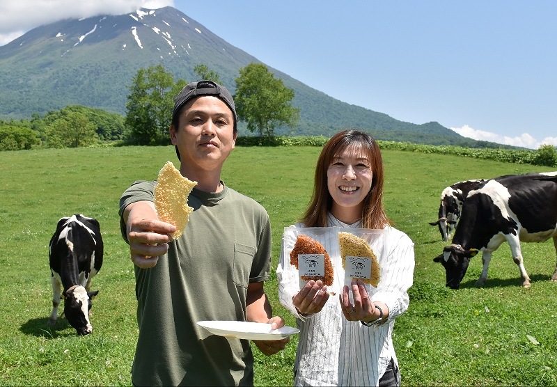 放牧地に立ち、レシピを受け継いだクリスピーチーズを手にする鈴木登さん(左)、塁さん夫妻