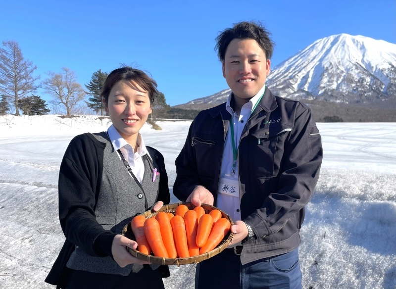 羊蹄山をバックに雪下ニンジンのおいしさをPRするJAようていの篠原佳夏さん(左)と新谷一馬さん